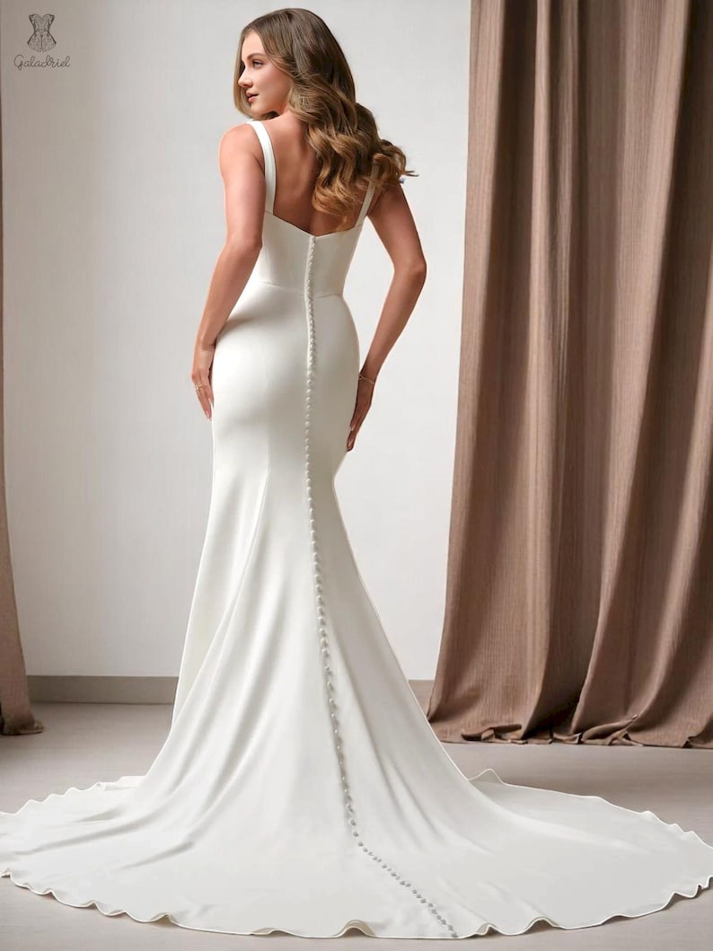K&ouml;nnte beinhalten: Ein wei&szlig;es Brautkleid mit einer figurbetonten Silhouette und einer langen Schleppe. Das Kleid hat einen quadratischen Ausschnitt, Spaghettitr&auml;ger und eine Reihe von Kn&ouml;pfen am R&uuml;cken. Das Kleid ist aus einem glatten, flie&szlig;enden Stoff gefertigt.