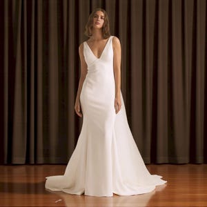 Puede incluir: Un vestido de novia blanco con escote en V y falda de sirena. El vestido tiene un corpi&ntilde;o ajustado y una larga cola. La modelo est&aacute; de pie frente a una cortina marr&oacute;n.