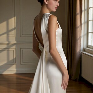 K&ouml;nnte beinhalten: Ein &auml;rmelloses, elfenbeinfarbenes Brautkleid mit tiefem R&uuml;ckenausschnitt und flie&szlig;ender Schleppe. Das Kleid ist aus einem glatten, satinartigen Stoff gefertigt und hat ein schlichtes, elegantes Design. Geeignet f&uuml;r Hochzeiten und formelle Anl&auml;sse.