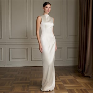 K&ouml;nnte beinhalten: &Auml;rmelloses, elfenbeinfarbenes Kleid mit hohem Ausschnitt. Das Kleid hat eine figurbetonte Silhouette, die bis zum Boden reicht. Das Model steht in einem Raum mit Holzboden und einem Fenster mit braunen Vorh&auml;ngen.