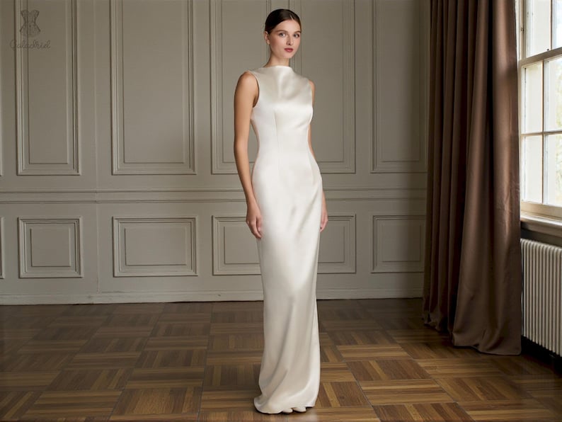 K&ouml;nnte beinhalten: &Auml;rmelloses, elfenbeinfarbenes Kleid mit hohem Ausschnitt. Das Kleid hat eine figurbetonte Silhouette, die bis zum Boden reicht. Das Model steht in einem Raum mit Holzboden und einem Fenster mit braunen Vorh&auml;ngen.