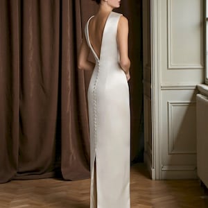 K&ouml;nnte beinhalten: Ein &auml;rmelloses, elfenbeinfarbenes Kleid mit tiefem V-Ausschnitt am R&uuml;cken und hohem Schlitz. Das Kleid hat eine Reihe kleiner Kn&ouml;pfe am R&uuml;cken und einen bodenlangen Rock. Das Model steht in einem Raum mit braunen Vorh&auml;ngen und Holzboden.
