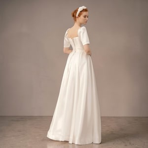 Puede incluir: Un vestido de novia blanco con escote cuadrado y mangas cortas. El vestido tiene un corpiño ajustado y una falda amplia y fluida. La modelo lleva una diadema blanca. El vestido está hecho de una tela lisa, posiblemente satén.