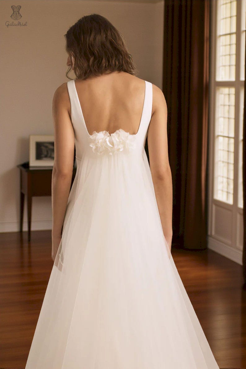 Puede incluir: Vestido de novia blanco con escote cuadrado y falda de tul fluida. La espalda presenta un adorno floral decorativo. El vestido es sencillo y elegante, adecuado para una boda o una ocasi&oacute;n especial.