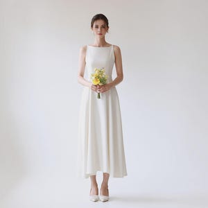 May include: A sleeveless, floor-length white dress with a boat neckline and a fitted bodice that flares into a full skirt. The dress is paired with pointed-toe white heels and a small bouquet of yellow and white flowers held at the waist.