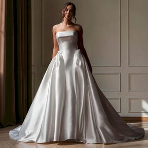 May include: A floor-length white satin gown featuring a strapless bodice with a straight neckline and a full, voluminous skirt. The dress includes decorative bow accents at the waistline, creating a structured silhouette with a smooth, lustrous finish.