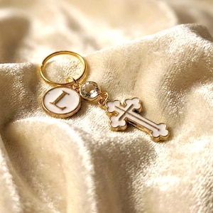 Può includere: Un portachiavi color oro con un ciondolo bianco a forma di lettera "L" e un ciondolo a forma di croce bianca. I ciondoli sono collegati da un anello dorato e un cristallo trasparente. Lo sfondo è un tessuto strutturato color crema.