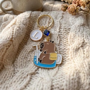 Può includere: Un portachiavi dorato con un animale marrone dei cartoni animati in una tazza blu, un cerchio bianco con la lettera "L" e una gemma blu. Il portachiavi è su una coperta a maglia a trecce color crema, con una tazza di tè e un libro aperto sullo sfondo.