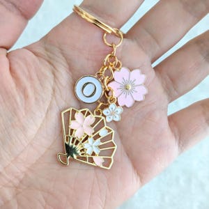 以下が含まれることがあります： ピンクと白の扇子のチャーム、ピンクの花のチャーム、白い花のチャーム、そして「O」の文字が書かれた白い丸いチャームが付いたゴールドのキーホルダーです。