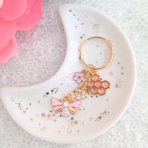 以下が含まれることがあります： ピンクと白の蜂のチャーム、ピンクの花のチャーム、ハニカムのチャームが付いたゴールドのキーチェーン。キーチェーンは、ゴールドの斑点のある白いセラミックの皿の上に置かれています。