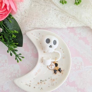 May include: A white ceramic crescent moon trinket dish with gold speckles. It features a white skull, a small white cylinder, and a charm with a black bow and a gold pendant. The dish is on a pink and white lace surface.