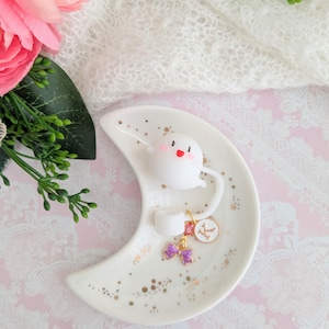 May include: A white ceramic crescent-shaped trinket dish with gold speckles. It features a cartoon ghost figure and a charm with a purple bow and the letter "K". The dish is on a pink and white lace surface.