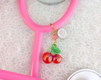 May include: A pink stethoscope with a gold charm attached. The charm features a pair of red cherries with green leaves, a pink crystal, and a white disc with the letter "B" in gold.