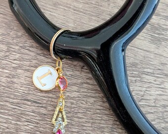 May include: A black stethoscope with a gold charm. The charm features a white circle with the letter "L", a red gem, and a gold and crystal pendant. The charm is attached to the stethoscope with a gold ring.