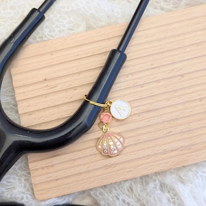 May include: A stethoscope charm featuring a gold ring with a white circular pendant with the letter "A", two pink gems, and a pink seashell charm with clear rhinestones. The stethoscope is black.