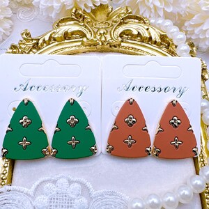 May include: Two pairs of triangle-shaped earrings with a geometric pattern. One pair is green and the other is orange. The earrings are on a white card with the word "Accessory" printed on it.