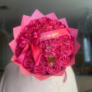 May include: A bouquet of bright pink satin roses, wrapped in pink paper, with a pink ribbon and a small perfume bottle. The ribbon has the text "I love you" in gold lettering. A romantic gift.