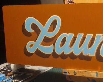 Op de afbeelding: Een bruin rechthoekig bord met het woord "Laundrette" in blauwe en witte cursieve letters.