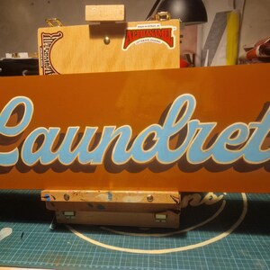 May include: A brown sign with the word "Laundrette" written in blue cursive lettering.