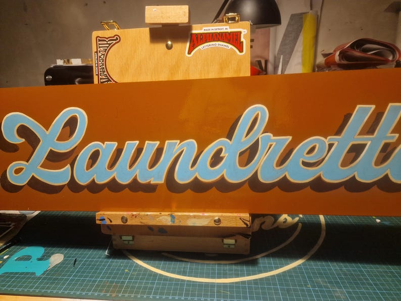May include: A brown sign with the word "Laundrette" written in blue cursive lettering.