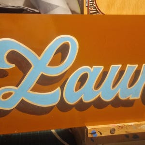May include: A brown wooden sign with the word "Laundry" painted in blue and white cursive lettering.