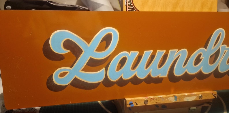 May include: A brown wooden sign with the word "Laundry" painted in blue and white cursive lettering.