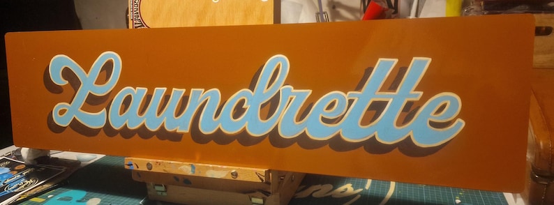 May include: A brown rectangular sign with the word "Laundrette" written in blue and white cursive letters.