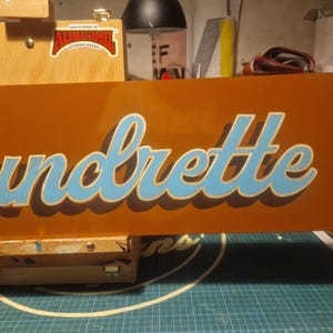 May include: A close-up of a wooden sign with the word "Laundrette" painted in blue and white lettering on an orange background.