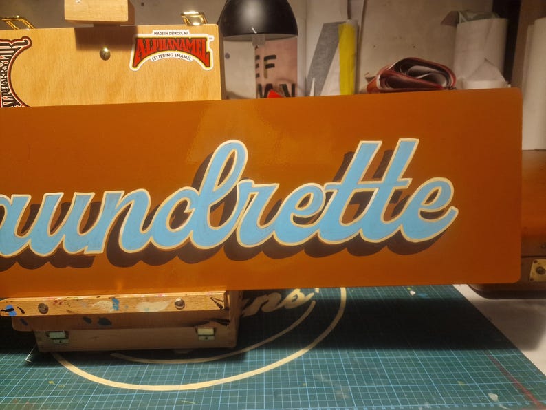 May include: A close-up of a wooden sign with the word "Laundrette" painted in blue and white lettering on an orange background.