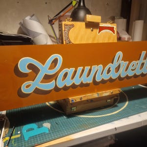 May include: A brown wooden sign with the word "Laundrette" painted in blue and white cursive lettering.
