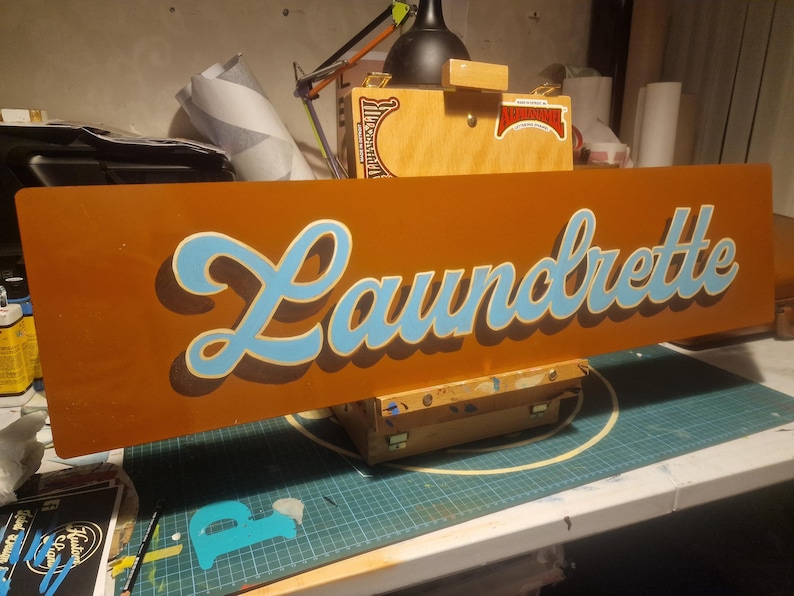 May include: A brown wooden sign with the word "Laundrette" painted in blue and white cursive lettering.