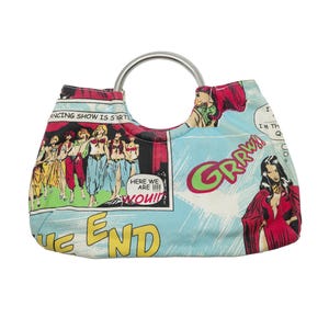 Puede incluir: Bolso con diseño de cómic y asa plateada. El bolso presenta paneles de cómic con ilustraciones de mujeres, bocadillos y las palabras "GRRWA" y "THE END". El fondo es azul claro.