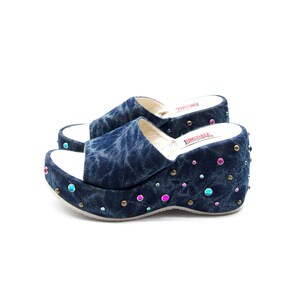 Puede incluir: Sandalias de plataforma vaqueras con suela blanca y parte superior de mezclilla azul. Las sandalias están decoradas con pedrería de colores y pequeños tachuelas doradas. El nombre de la marca "TUNSPALE" está impreso en la plantilla.