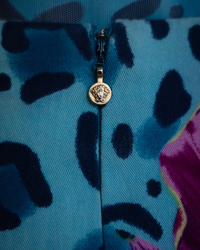 K&ouml;nnte beinhalten: Nahaufnahme eines Stoffes mit blauem und schwarzem Muster und einem goldenen Rei&szlig;verschluss-Zipper mit einem Kopf mit Krone. Der Rei&szlig;verschluss-Zipper ist rund.