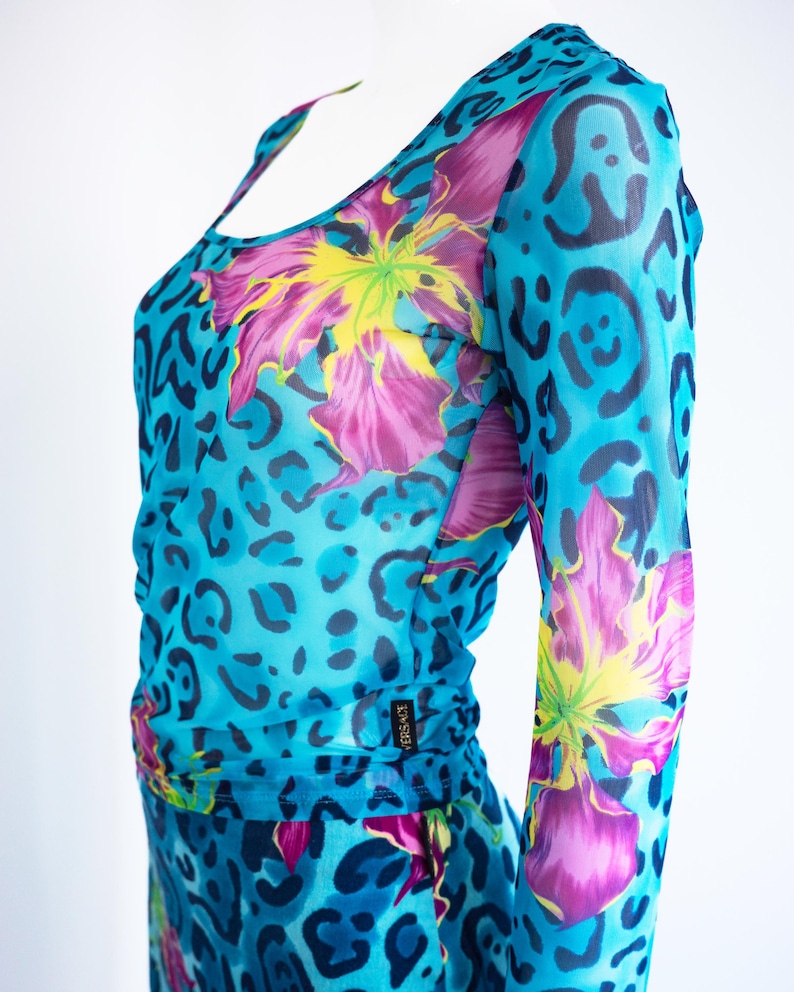 K&ouml;nnte beinhalten: Ein blaues Netz-Oberteil mit Blumen- und Leopardenmuster. Das Oberteil hat lange &Auml;rmel und einen runden Ausschnitt. Das Label "VERSACE" ist an der Seite sichtbar.