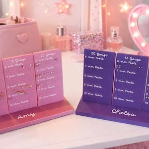 May include: Two earring display stands, one pink and one purple, with various earrings. The pink stand is labeled "Amy" and the purple stand is labeled "Chelsa". Each stand has multiple rows of earrings with gauge and post size information.