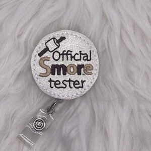 May include: A round, white badge with the text "Official S'more tester" embroidered in black and brown. The badge features a graphic of a marshmallow on a stick and is attached to a clear plastic clip.
