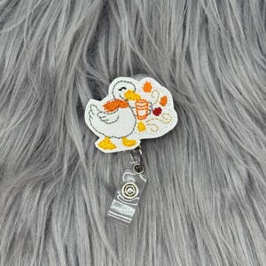 May include: A white embroidered duck badge reel featuring an orange scarf and holding an orange beverage cup. The duck is surrounded by small embroidered autumn leaves in shades of orange, yellow, and red, attached to a clear plastic strap.
