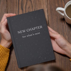 May include: A dark gray journal held by a person, with the words "NEW CHAPTER" and "for what's next" printed in white. A white mug with a beverage sits on a wooden surface.