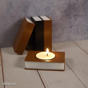 May include: A brown book-shaped candle holder with a lit candle inside. The candle holder is made to look like a book with white pages and a brown cover. Two other book-shaped objects are behind the candle holder.