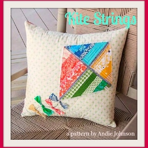 Può includere: Un cuscino quadrato color crema con un design a patchwork di aquilone nei toni del rosso, blu, verde e giallo. Il cuscino ha un motivo sottile e code di aquilone decorative. Il testo sul cuscino recita "Kite Strings" e "a pattern by Andie Johnson."