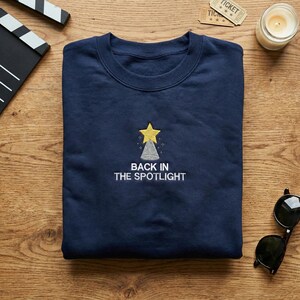 May include: A navy blue sweatshirt featuring a gold star and spotlight design, with the text "BACK IN THE SPOTLIGHT" embroidered below. The sweatshirt is folded on a wooden surface, with a film clapper, sunglasses, and a candle visible.