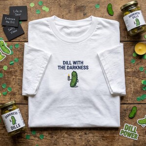 May include: White t-shirt featuring the phrase "DILL WITH THE DARKNESS" and a cartoon pickle with a candle. The image also shows jars of pickles, stickers, and small black signs with text.