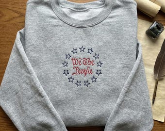 May include: A heather gray sweatshirt with a crew neck. The front features an embroidered design of a circle of blue stars surrounding the words "We The People" in red and blue script. The sweatshirt is folded on a wooden surface.