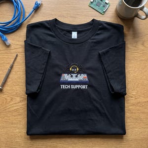 May include: A black short-sleeve t-shirt featuring an embroidered graphic of a spider wearing yellow headphones and a microphone while typing on a computer keyboard. Below the graphic, the text TECH SUPPORT is embroidered in white.