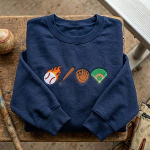 May include: Navy blue sweatshirt with embroidered baseball-themed designs. Features a baseball on fire, a wooden bat, a baseball glove, and a baseball field. The sweatshirt is made of a soft material.