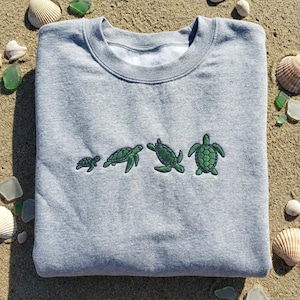 Könnte beinhalten: Ein graues Sweatshirt mit einer Stickerei von vier grünen Schildkröten, die in der Größe zunehmen. Das Sweatshirt wird auf Sand präsentiert, umgeben von Muscheln und Meeresglas.