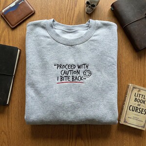 May include: A heather gray sweatshirt with the embroidered text "PROCEED WITH CAUTION I BITE BACK". The text is in black with a red underline. The sweatshirt is folded on a wooden surface, surrounded by accessories.