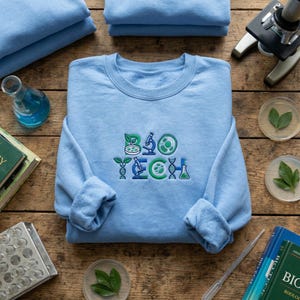 May include: A light blue sweatshirt with the embroidered text "BIO TECH" in green and blue. The design includes images of leaves, a microscope, and a DNA strand. The sweatshirt is folded on a wooden surface with lab equipment.