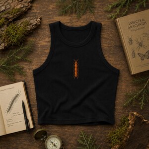 Könnte beinhalten: Ein schwarzes, geripptes Tanktop mit einer kleinen, mittig platzierten orangefarbenen Tausendfüßler-Grafik. Das Kleidungsstück hat einen hohen, runden Ausschnitt und ein ärmelloses Design, das eine schlichte, körpernahe Silhouette zeigt.